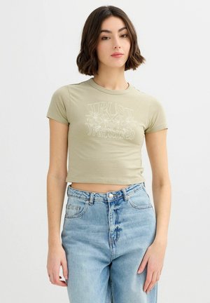 Joven con cabello corto y oscuro que lleva un top corto verde claro con el texto "Confía en tus sentidos" y unos vaqueros de talle alto azul claro.