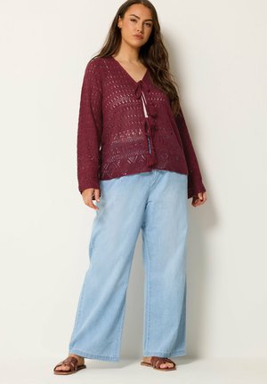 Vrouw die een bordeauxgebreid vest met kwastjes draagt, lichtblauwe wijde jeans en bruine sandalen met open teen, staand tegen een effen achtergrond.
