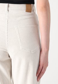 Personne portant un pantalon en velours côtelé blanc taille haute avec poche arrière et étiquette de marque en cuir marron, bras détendu sur le côté.