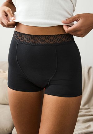 Close-up van een persoon die zwart boxershort-ondergoed met een kanten tailleband draagt en een witte geribbelde tanktop optilt.