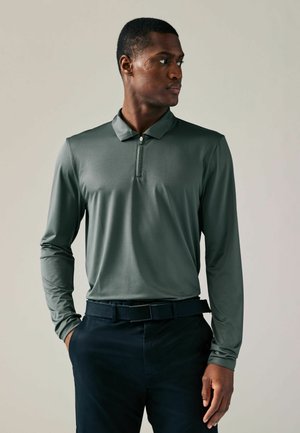 Long-sleeve performance polo in dark green, featuring a half-zip collar, textured fabric, and paired with dark navy trousers and a black belt.