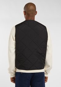 Chaleco acolchado negro con un patrón de diamantes, que presenta un cuello alto y sisas, superpuesto a un suéter de color crema.