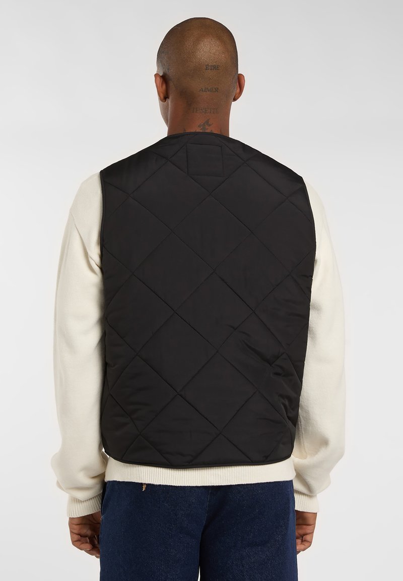 Sort quilted vest med diamantmønster, der har høj krave og ærmegab, lagdelt over en cremefarvet sweater.