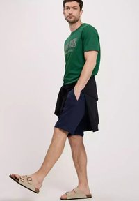 Zielony graficzny T-shirt, granatowe szorty, czarny sweter związany w talii, beżowe sandały. Strój charakteryzuje się swobodnym, luźnym stylem i materiałami.