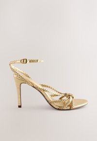 Sandale à talon haut dorée avec des lanières tressées, une boucle à la cheville et une finition métallique lisse. Design élégant à bout ouvert et talon fin.