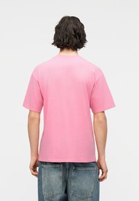 Άτ ομο με μεσαίου μήκους σκούρα μαλλιά, που φορά ένα απλό ροζ κοντομάνικο μπλουζάκι και μπλε τζιν, με την πλάτη προς τα εμπρός σε λευκό φόντο.