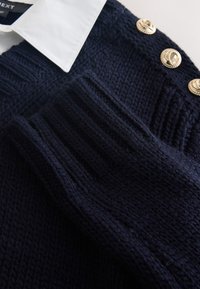 Mørkeblå chunky strikket sweater med en hvid skjorte under og tre gyldne dekorative knapper på skulderen.