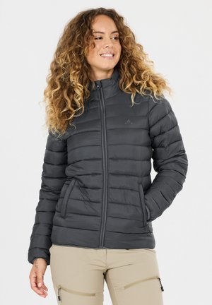 Femme aux cheveux bouclés portant une doudoune noire zippée et un pantalon beige avec des poches zippées, souriante et regardant vers sa gauche.