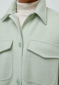 Giacca camicia in lana di colore verde chiaro, con colletto, due tasche sul petto e bottoni abbinati. Texture morbida e design informale.