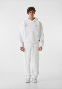 Hvit hettegenser med rosa brodert tekst, med en frontlomme og ribbestrikkede mansjetter; kombinert med matchende hvite joggebukser og sneakers.