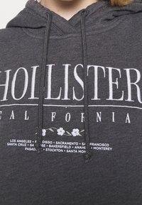 Grå huvtröja med vit text "HOLLISTER" och "CALIFORNIA", med blommiga detaljer och en huva med justerbara snören.