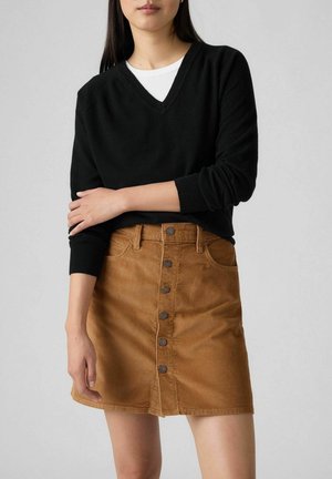 Femme portant un pull noir à manches longues et col en V par-dessus une chemise blanche et une jupe en velours côtelé marron avec des boutons sur le devant, debout devant un fond uni.