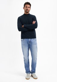 Donkerblauwe gebreide trui met een kwart-rits kraag, gecombineerd met lichtblauwe jeans en grijze sneakers. Ribgebreide manchetten en tailleband.