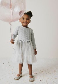 Lichtgrijze kabelgebreide trui met lange mouwen, gecombineerd met een witte tule jurk versierd met zilveren pailletten, en glinsterende zilveren schoenen. Het kind houdt een grote ballon vast.
