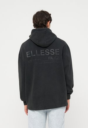 Persona con sudadera negra con capucha con "ELLESSE" y logo impreso en la espalda, combinada con pantalones gris claro moteados.