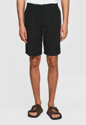 Mężczyzna noszący czarne szorty ze ściągaczem i podwiniętymi nogawkami oraz brązowe sandały z podwójnym paskiem, stojący na jednolitym tle.