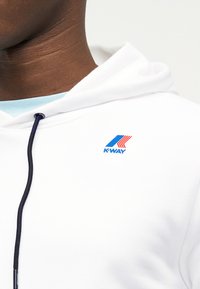 Κοντινό πλάνο ατόμου που φοράει λευκό φούτερ K-Way με λογότυπο μπλε και κόκκινο στο στήθος και μαύρο κορδόνι στη κουκούλα.