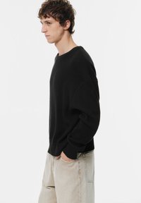 Maglione nero a coste con scollatura rotonda e maniche lunghe, abbinato a pantaloni beige chiaro. Il tessuto presenta una superficie testurizzata.