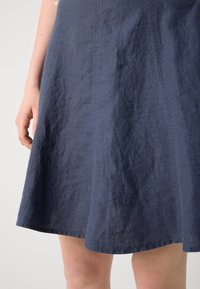 Gros plan sur une personne portant une jupe texturée bleu foncé arrivant au genou, avec des coutures verticales visibles et une main reposant sur le côté.
