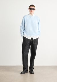 Sudadera azul claro de corte holgado, sobre una camisa blanca. Pantalones de denim gris oscuro. Zapatos negros y gafas de sol completan el atuendo.