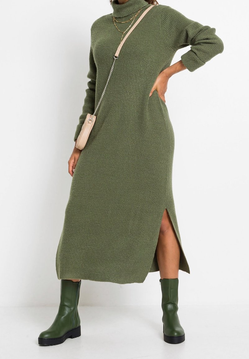 Femme portant une robe en maille côtelée verte à col roulé avec une fente latérale, des bottes vertes mi-mollet, des chaînes dorées superposées et un petit sac beige porté en bandoulière.