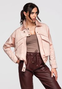 Femme aux cheveux foncés portant des boucles d’oreilles créoles dorées, une veste oversize rose clair, un top taupe et un pantalon en cuir bordeaux, posant avec la main sur la hanche.
