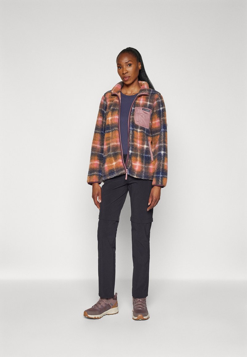 Veste en tartan flou dans des teintes d'orange, de bleu et de jaune avec une poche zippée rose ; associée à un pantalon sombre et des baskets violettes.