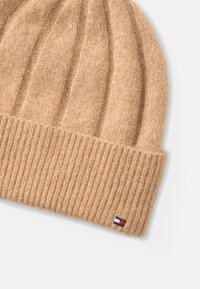 Knit beanie in light tan wool blend, featuring ribbed texture and a folded cuff. Small logo tag with red, white, and blue accent.