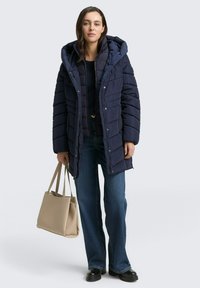 Granatowa kurtka puchowa z kapturem, pikowany design i zamek błyskawiczny. W połączeniu z czarnym topem, niebieskimi dzwonami i beżową torbą.