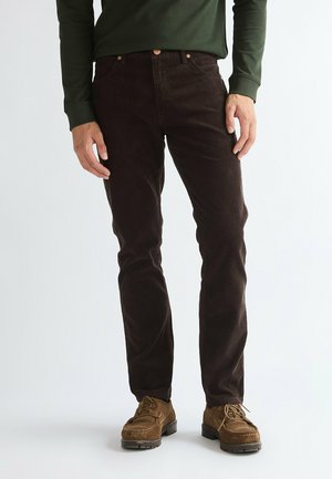 Uomo che indossa pantaloni di velluto a coste marrone scuro, scarpe marroni con lacci e una camicia verde scuro a maniche lunghe, in piedi davanti a uno sfondo semplice.