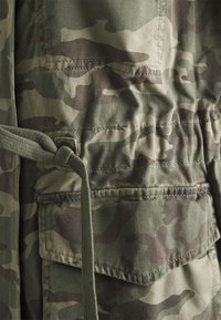 Camouflagepatroon stof in schakeringen van groen en bruin, met een trekkoord in de taille en een patch pocket detail aan de zijkant.
