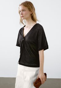 Svart kortärmad cardigan med djup V-ringning och knappslutning fram. Lätt, något genomskinligt tyg i kombination med en texturerad brun clutch.