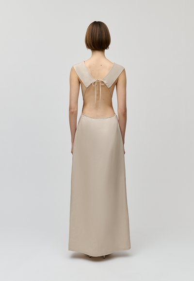 Abito maxi beige con schiena a V, spalline larghe e chiusura con lacci. Texture liscia con un design fluido, caratterizzato da una profonda schiena aperta.