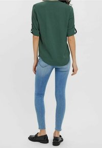Chemise verte à manches retroussées, avec un dos long, associée à un jean skinny bleu clair et des chaussures noires. Le tissu semble lisse et léger.