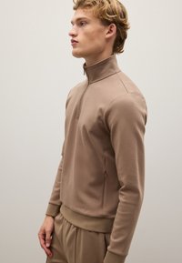 Brauner Reißverschluss-Pullover aus weichem, glattem Material. Verfügt über einen hohen Kragen, gerippte Bündchen und ein tailliertes Design. Seitentaschen sichtbar.