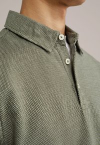 Cămașă verde texturată cu guler ascuțit, având butoane albe și un model țesut, prezentând un material subtil, ușor.