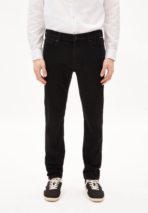 Mann trägt schwarze Slim-Fit-Jeans, weißes Hemd mit Knöpfen und schwarze Sneakers mit weißen Schnürsenkeln, steht mit entspannten Händen an den Seiten.