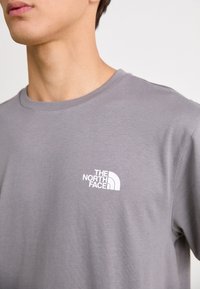 Szara koszulka z krótkim rękawem wykonana z bawełny, z białym logo "The North Face" na lewej części klatki piersiowej. Gładka faktura, dekolt w serek.