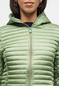 Γυναίκα με ανοιχτοπράσινο καπιτονέ μπουφάν κλειστό με φερμουάρ, φοράει χρυσούς κρίκους στα αυτιά κα ι μαύρο τοπ από κάτω.