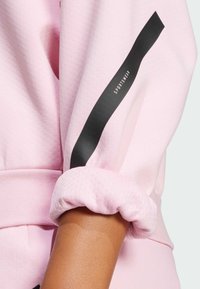 Soft pink sportswear top with a textured fabric, featuring a black stripe and logo on the sleeve. Cuffed sleeves add detail.
