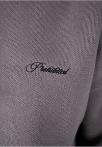 Gros plan d’un tissu tricot gris avec "Prediction" en cursive noire brodé dessus.