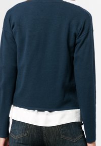 Navy textured sweater with a rounded hem, layered over a white shirt. Long sleeves with a relaxed fit. jeans visible at the waist.