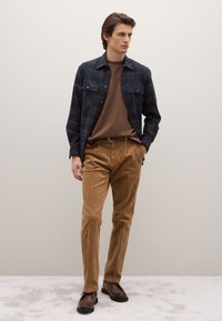 Camisa de cuadros azul marino y verde con botones, superpuesta sobre una camiseta marrón, combinada con pantalones de pana color beige y zapatos marrones. Diseño y texturas simples.