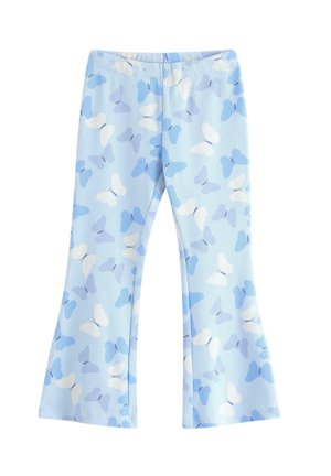 Pige smiler med hænderne på hofterne, iført hvid skjorte med blå sommerfugleapplikationer, lyseblå bukser med sommerfuglemønster og sneakers.