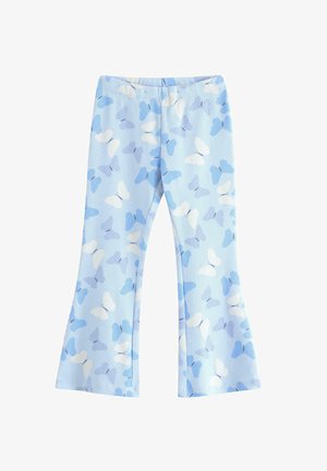 Pige smiler med hænderne på hofterne, iført hvid skjorte med blå sommerfugleapplikationer, lyseblå bukser med sommerfuglemønster og sneakers.