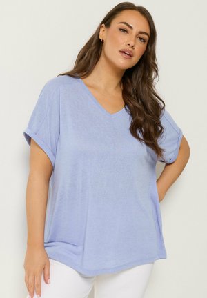 Donna con lunghi capelli castani che indossa una maglietta azzurro chiaro con scollo a V e maniche corte e pantaloni bianchi, posando con una mano sui fianchi.