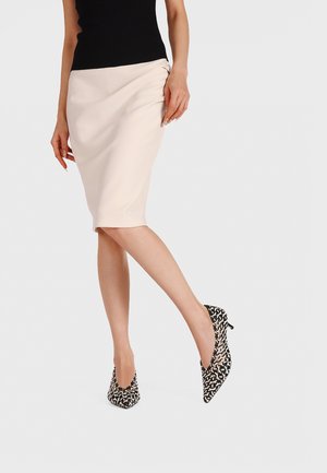 Femme portant une jupe crayon beige longueur genou, un haut noir sans manches, et des escarpins à bouts pointus noir et blanc avec talons petits.