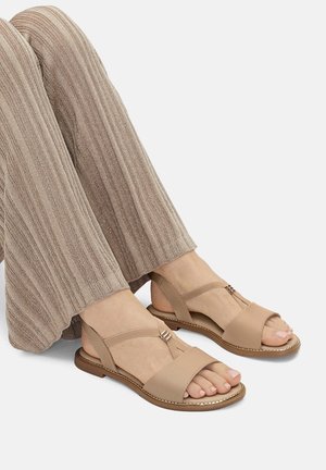 Beżowe sandały z odkrytymi palcami i paskiem wokół kostki, z T-kształtnym wzorem i metalicznymi akcentami. Noszone z teksturowanymi, jasnobrązowymi spodniami o szerokich nogawkach.