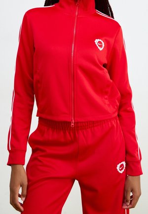 Vrouw draagt een felrode jas met ritssluiting en bijpassende hoge taille broek met witte strepen en witte logopatches op borst en dij.
