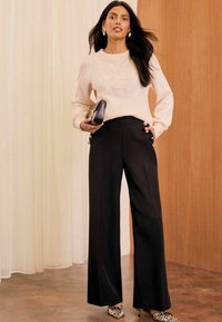 Gaiši rozā adīta džemperis ar tekstūras detaļām, apvienots ar augsto jostasvietu melniem platām biksēm ar zelta pogām un ar dzīvnieku rakstu apaviem.